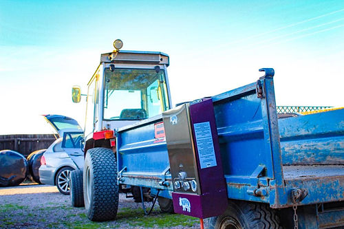 Tractor with a blue trailer attached, parked on a grassy area with cars in the background.