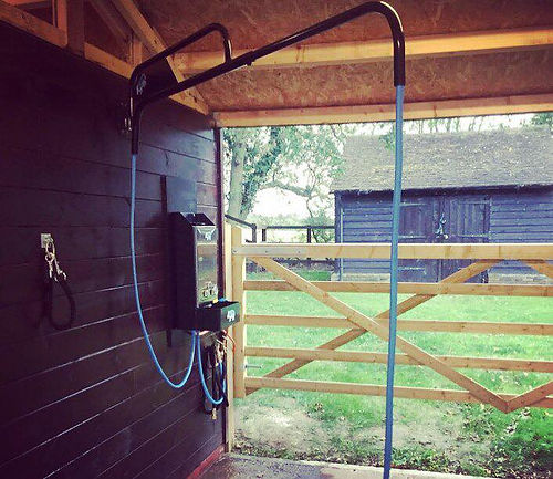 Outdoor shower with hose attached to a wooden building, with a fenced area and trees in the background.