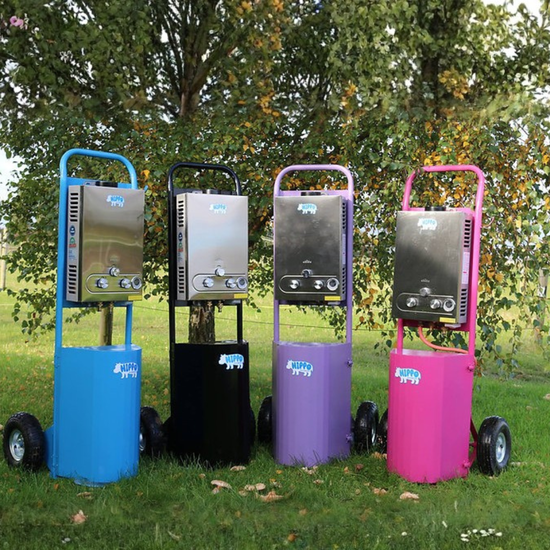Four portable gas showers on wheels with colorful stands in a grassy area with trees.