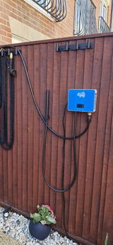 A blue electric shower wall box mounted on a brown wooden fence, with a black hose attached. The scene is set outdoors in a backyard with a brick wall and a small potted plant at the bottom.
